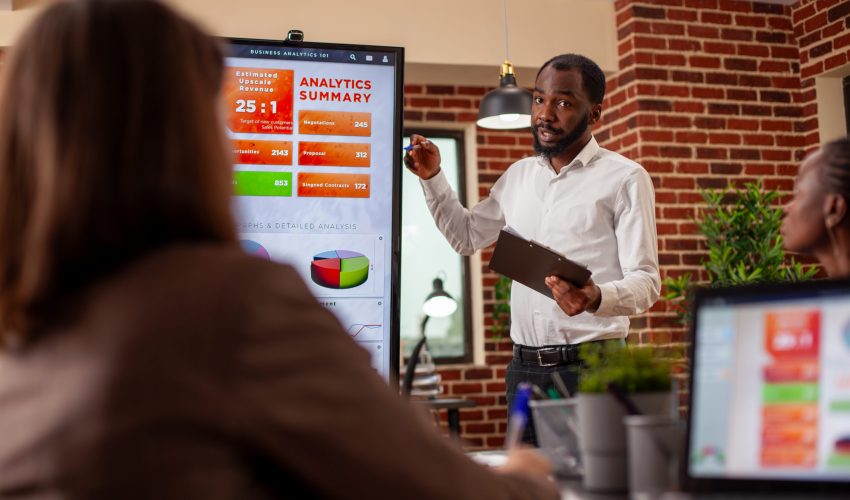Businessman with clipboard in hand, using digital monitor to explain analytics summary in meeting. African american entrepreneur doing project presentation, planning marketing strategy with his team.